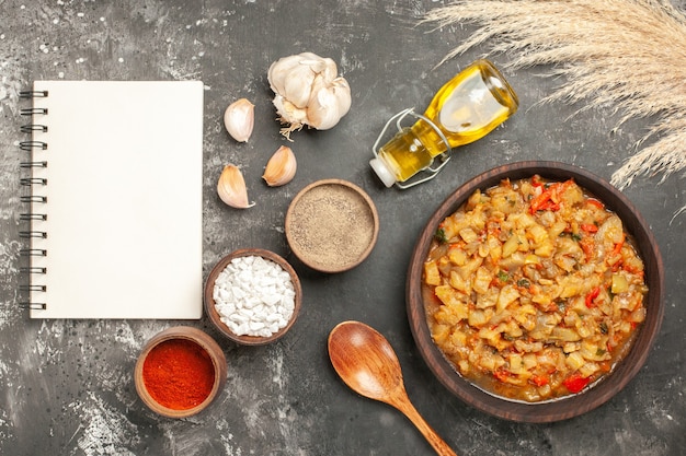 Vista dall'alto di insalata di melanzane arrosto in una ciotola, bottiglia di olio, cucchiaio di legno e diverse spezie nel taccuino di ciotole sulla superficie scura