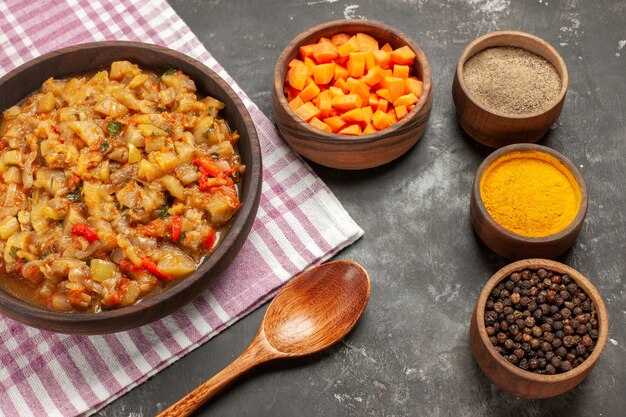 Vista dall'alto di insalata di melanzane arrosto in una ciotola ciotole con diverse spezie taglia cuccioli di carota cucchiaio di legno sulla superficie scura