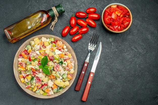 Vista dall'alto di insalata di verdure con bottiglia di olio di pomodori e posate sul lato su sfondo grigio scuro