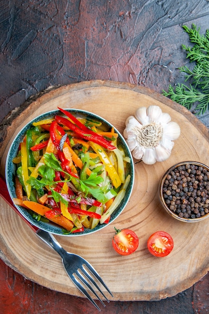 Vista dall'alto di insalata di verdure in ciotola forcella aglio pepe nero sul ramo di abete bordo rustico sulla tavola rosso scuro