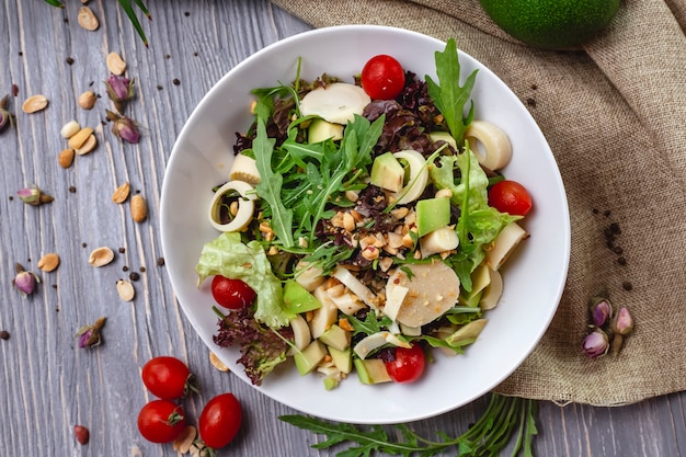 Vista dall'alto di insalata fresca con formaggio pomodorini avocado e arachidi tostate in una ciotola bianca su fondo rustico