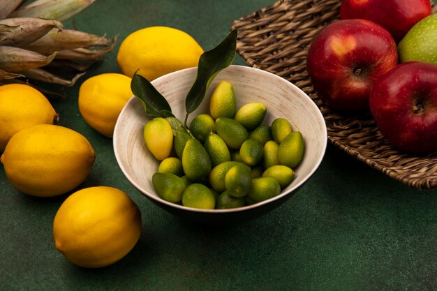Vista dall'alto di kinkan aromatici su una ciotola con mele rosse fresche su un vassoio di vimini con limoni isolato su una parete verde