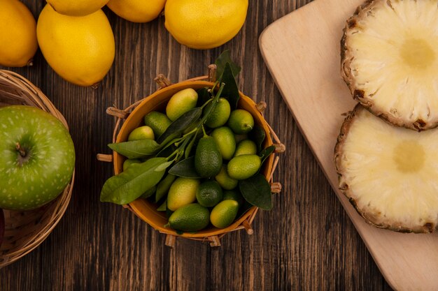Vista dall'alto di kinkan freschi su un secchio con ananas su una tavola da cucina in legno con mele sul secchio con limoni isolato su una parete in legno