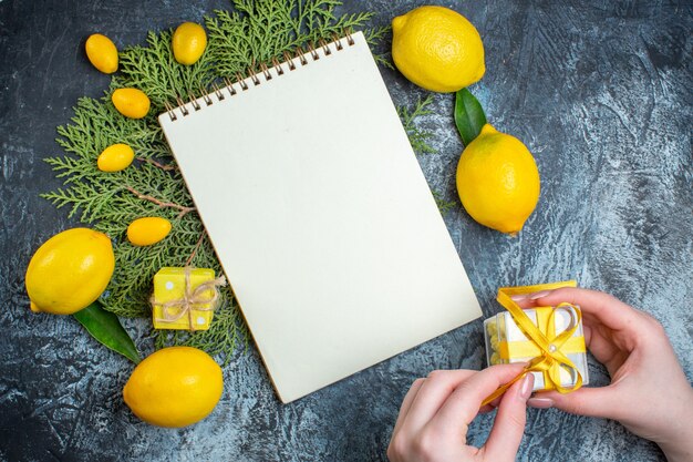 Vista dall'alto di limoni freschi con foglie e quaderno a spirale chiuso su rami di abete che aprono a mano una confezione regalo su sfondo scuro