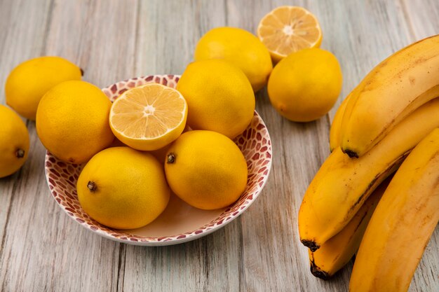 Vista dall'alto di limoni interi dalla pelle gialla su una ciotola con le banane isolate su un fondo di legno grigio