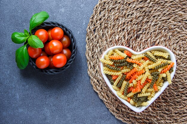 Vista dall'alto di maccheroni colorati pasta a forma di cuore ciotola con pomodori in una ciotola, foglie su sottopentola e superficie grigia. orizzontale