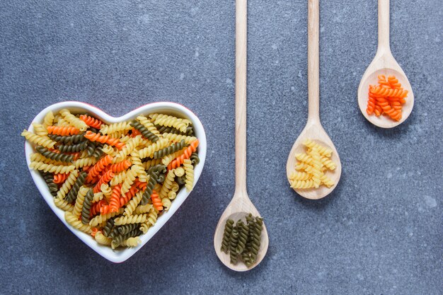 Vista dall'alto di maccheroni colorati pasta a forma di cuore ciotola e cucchiai sulla superficie grigia. orizzontale