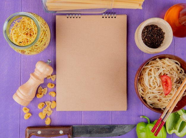 Vista dall'alto di maccheroni come spaghetti cotti e crudi farfalle pipe-rigate bucatini con coltello pepe nero burro pepe intorno a blocco note su sfondo viola con spazio di copia