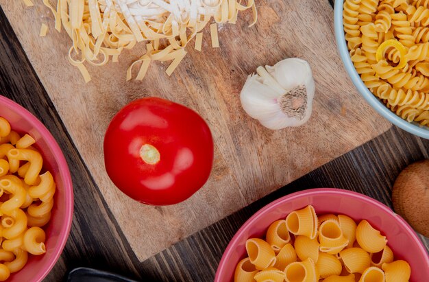 Vista dall'alto di maccheroni tagliatelle con farina aglio e pomodoro sul tagliere con altri tipi di pasta su legno