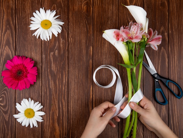 Vista dall'alto di mani che legano con un nastro un bouquet di fiori rosa e bianchi alstroemeria e calla su fondo in legno