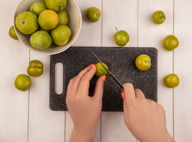 Vista dall'alto di mani femminili che tagliano prugne ciliegia verde su un tagliere di cucina con coltello con prugne ciliegia verde su una ciotola su un fondo di legno bianco