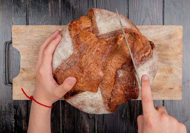 vista dall'alto di mani maschili taglio pane di pannocchia con coltello sul tagliere su fondo in legno