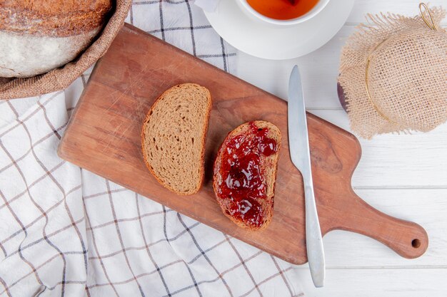 Vista dall'alto di marmellata di fragole spalmato sul pane di segale affettato con coltello sul tagliere e pannocchia sul panno plaid con tè e marmellata sul tavolo di legno