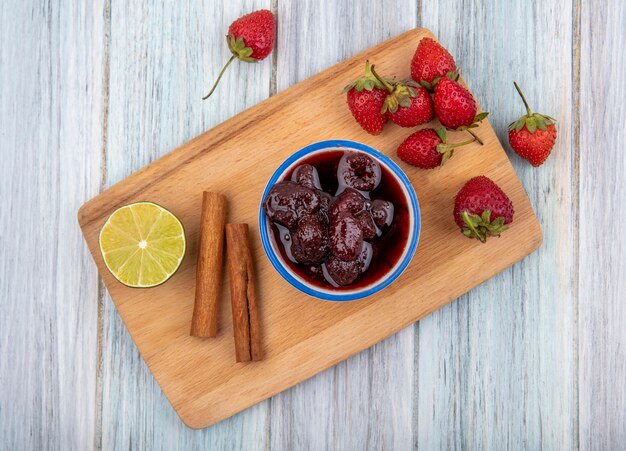 Vista dall'alto di marmellata di fragole su una ciotola blu su una tavola da cucina in legno con fragole fresche con calce con bastoncini di cannella su un fondo di legno grigio