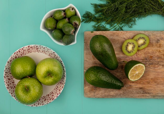 Vista dall'alto di mele verdi fresche su una ciotola con feijoas su una ciotola con avocado limette e kiwi su una tavola da cucina in legno su una parete blu