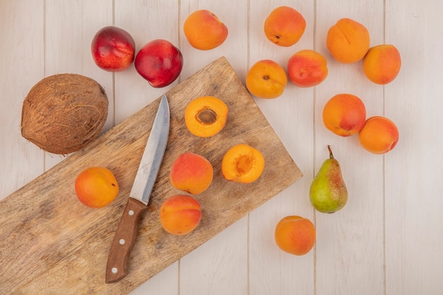 Vista dall'alto di metà tagliate e intere albicocche e coltello sul tagliere con motivo di frutta come pesche albicocche pera e cocco su sfondo di legno