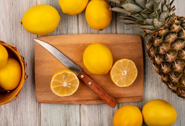 Vista dall'alto di mezze limoni freschi su una tavola da cucina in legno con coltello con limoni su un secchio con limoni e ananas isolato su una superficie di legno grigia