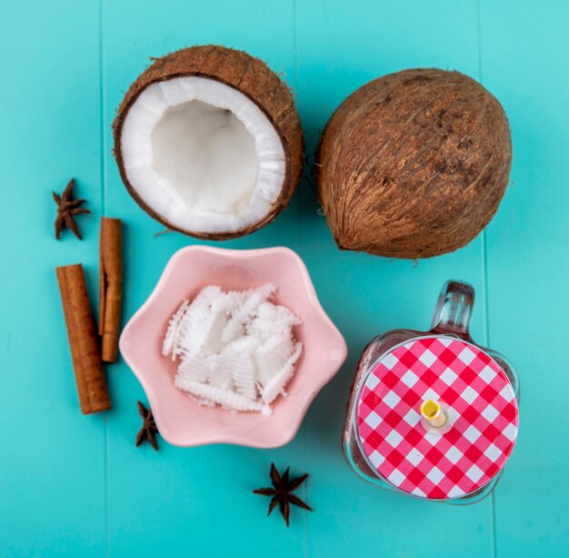 Vista dall'alto di noci di cocco dimezzate e intere con polpa di cocco in una ciotola rosa con succo di anice e cannella in vaso di vetro sulla superficie blu