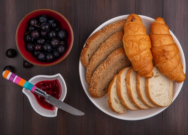 Vista dall'alto di pane come baguette di pannocchia marrone seminate a fette e croissant nel piatto e ciotole di marmellata di lamponi e prugnole con coltello su sfondo di legno