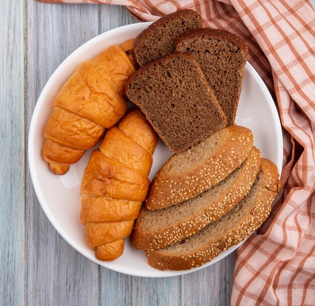 Vista dall'alto di pane come croissant a fette di segale e seminate pannocchia marrone nella piastra su plaid panno su sfondo di legno