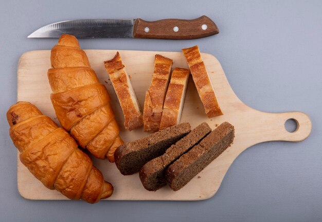 Vista dall'alto di pane come croissant di segale e baguette sul tagliere e coltello su sfondo grigio