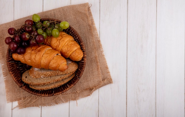 Vista dall'alto di pane come croissant e semi di pannocchia marrone fette di pane con uva nel cesto su tela di sacco su sfondo di legno con spazio di copia