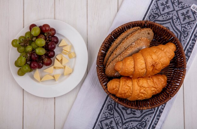 Vista dall'alto di pane come croissant e semi di pannocchia marrone fette di pane nel cestello su stoffa e piatto di uva e formaggio su sfondo di legno