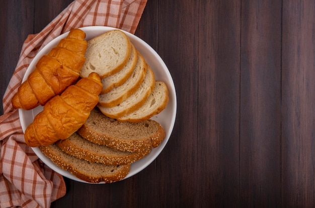 Vista dall'alto di pane come fette di pannocchia marrone con semi di baguette e croissant nella piastra sul panno plaid su sfondo di legno con spazio di copia