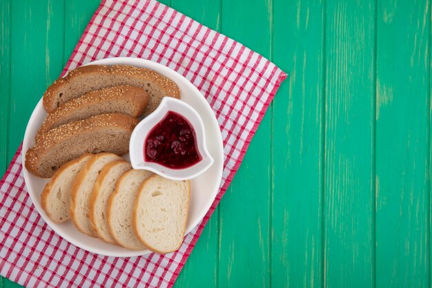 Vista dall'alto di pane come fette di pannocchia marrone seminato e baguette con una ciotola di marmellata di lamponi nella piastra su panno plaid su sfondo verde con spazio di copia