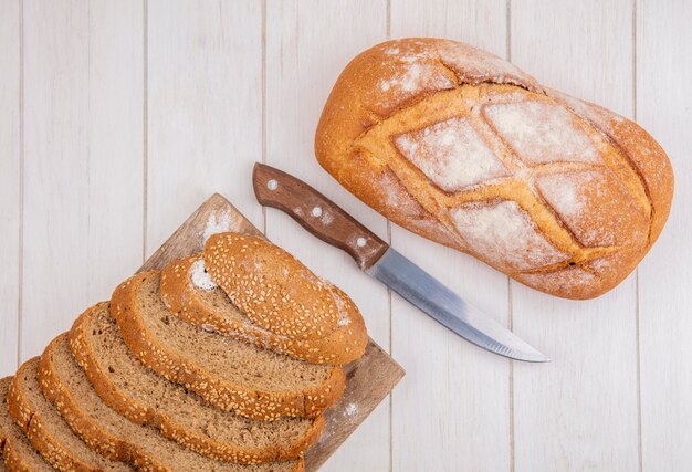 Vista dall'alto di pane come pannocchia con semi marrone a fette sul tagliere e pane croccante con coltello su sfondo di legno