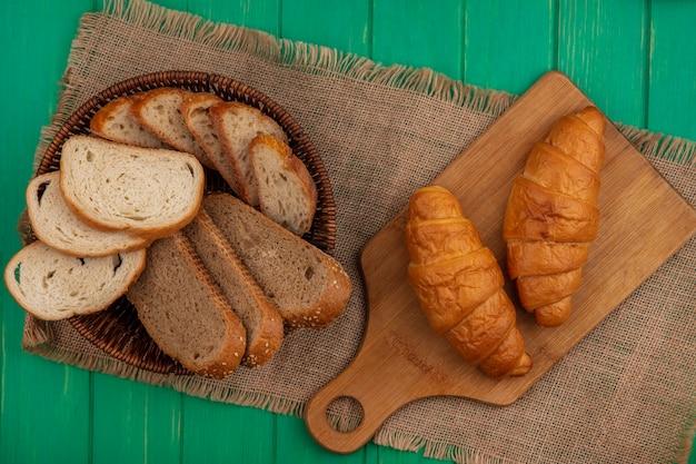 Vista dall'alto di pane come pannocchia marrone seminate e fette di baguette nel cestello e croissant sul tagliere su tela di sacco su sfondo verde