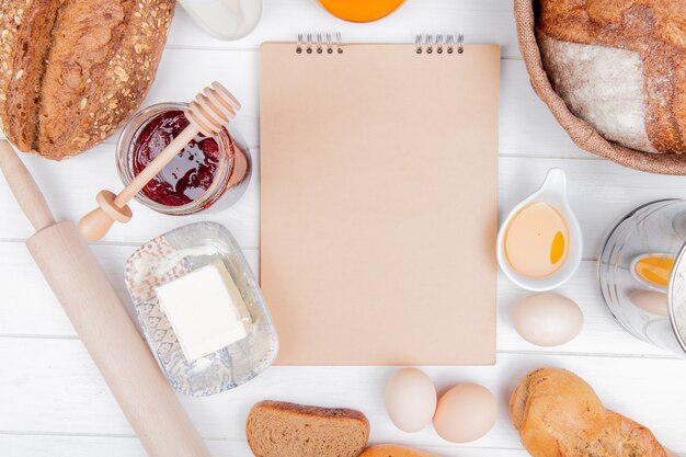 Vista dall'alto di pane come pannocchie di pane di segale baguette con semi e vietnamita con burro di uova marmellata mattarello e blocco note su fondo in legno con spazio di copia