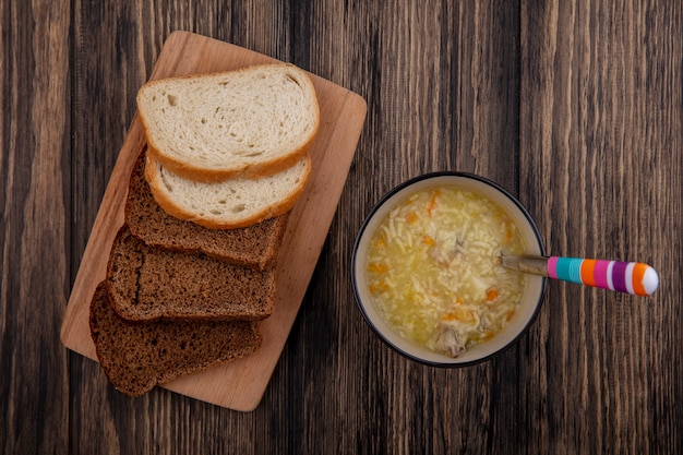 Vista dall'alto di pane come segale a fette e quelli bianchi sul tagliere e ciotola di zuppa di pollo orzo con cucchiaio su sfondo di legno