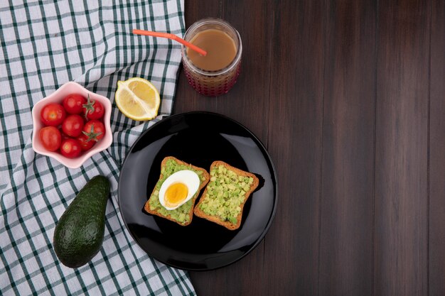 Vista dall'alto di pane tostato con polpe di avocado e uovo con pomodori su una ciotola rosa limone avocado su tovaglia a quadretti e superficie in legno