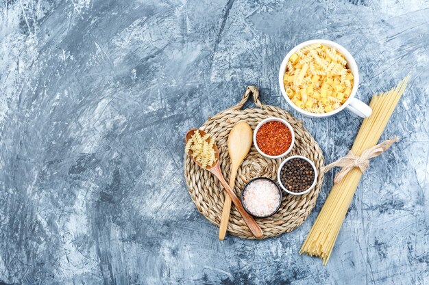 Vista dall'alto di pasta assortita in una ciotola con cucchiai di legno, spezie su intonaco grigio