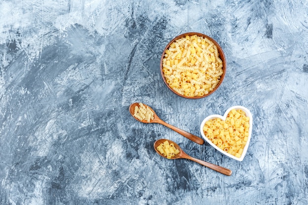 Vista dall'alto di pasta cruda in ciotole e cucchiai di legno su sfondo grigio intonaco. orizzontale