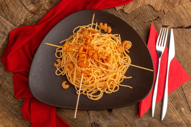 Vista dall'alto di pasta italiana cotta con gamberetti all'interno del piatto marrone con posate sulla superficie in legno
