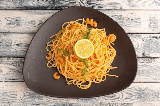 Vista dall'alto di pasta italiana cotta con gamberi verdi e limone all'interno del piatto marrone sulla superficie di legno grigia