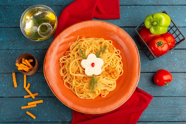 Vista dall'alto di pasta italiana cotta con verdure all'interno del piatto arancione con olio e verdure sulla superficie in legno blu