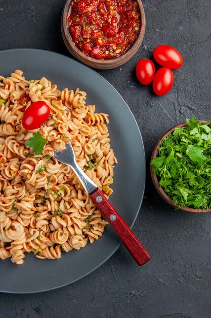 Vista dall'alto di pasta rotini con forchetta di pomodorini su piastra prezzemolo e salsa di pomodoro in ciotole pomodorini su superficie scura