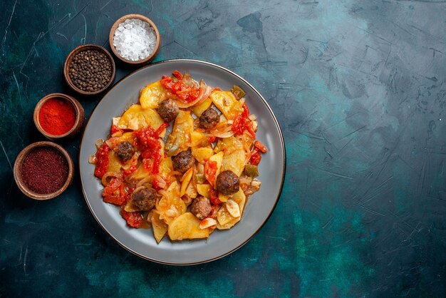 Vista dall'alto di patate al forno con polpette di carne e verdure insieme a condimenti su sfondo blu scuro.