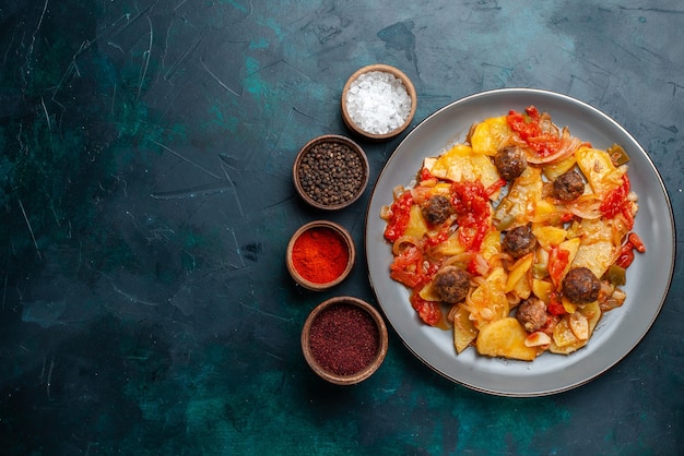 Vista dall'alto di patate al forno con polpette di carne e verdure insieme a condimenti sulla scrivania blu scuro.