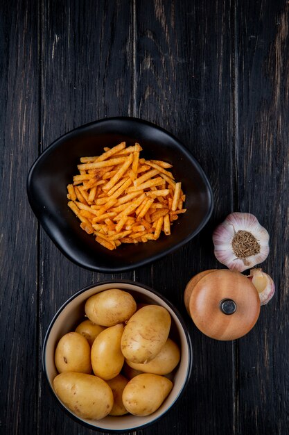 Vista dall'alto di patate in ciotole fritte e crude intere con sale e aglio su legno