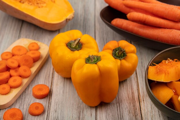 Vista dall'alto di peperoni arancioni con fette di zucca su una ciotola con carote tritate su una tavola da cucina in legno su una parete di legno grigia