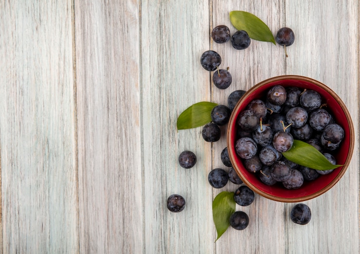 Foto gratuito vista dall'alto di piccole prugnole di frutta viola scuro acide su una ciotola con foglie su un fondo di legno grigio con spazio di copia