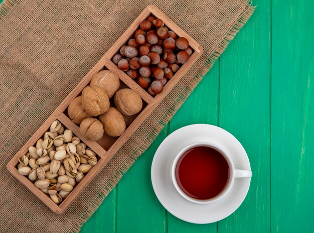 Vista dall'alto di pistacchi con nocciole e noci su un tovagliolo beige con una tazza di tè su una superficie verde