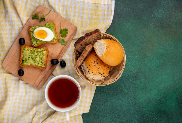 Vista dall'alto di polpe di avocado su pane tostato con uovo sulla cucina in legno bordo con un secchio di pane su giallo tovaglia a quadretti e gre