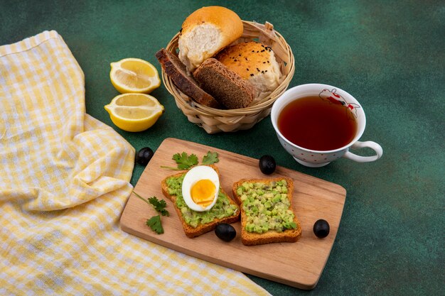 Vista dall'alto di polpe di avocado su pane tostato con uovo sulla tavola da cucina in legno con una tazza di tè e un secchio di pane su gre