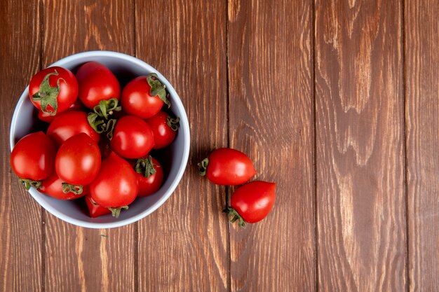 Vista dall'alto di pomodori in una ciotola con altri sul lato sinistro e superficie in legno con spazio di copia