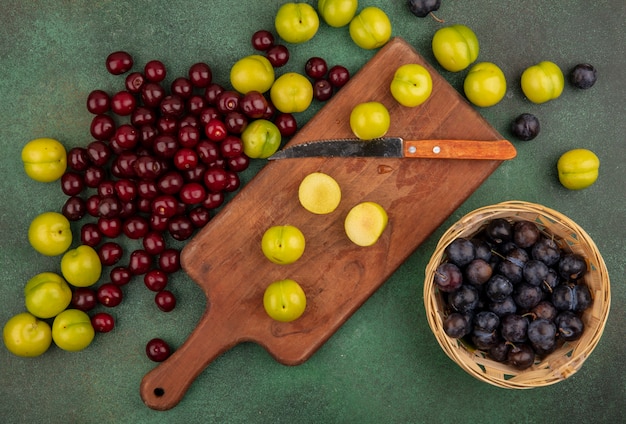 Vista dall'alto di prugne ciliegie verdi fresche su una tavola da cucina in legno con coltello con ciliegie rosse con prugnole su un secchio su sfondo verde